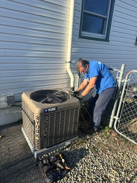 HVAC technician in a blue shirt repairing a York outdoor air conditioner next to the house exterior, on a gravel area with tools nearby.