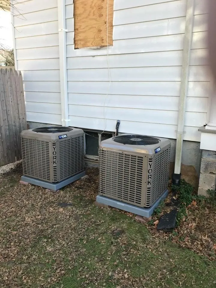Two outdoor York air conditioning condensers on concrete pads beside a house, with a plywood-covered window above.