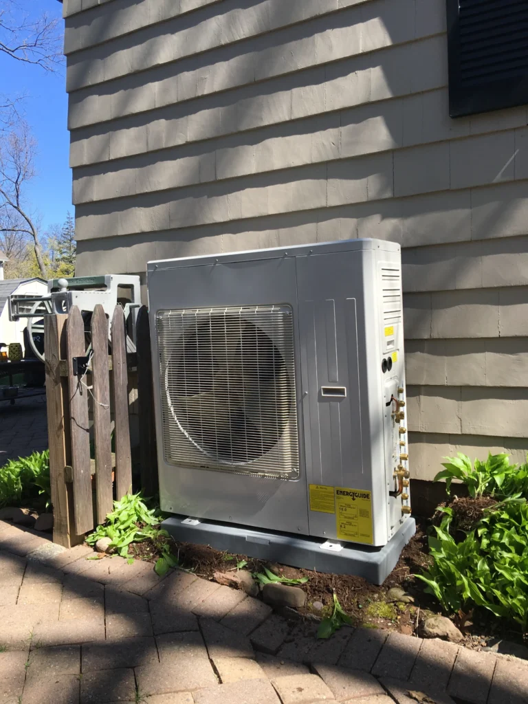 Outdoor HVAC condenser unit beside a beige siding house, next to a wooden fence and landscaped plants in a sunny yard.